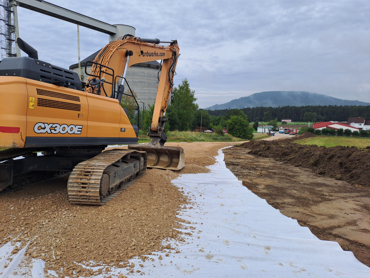 Baustelle mit Bagger auf einem mit Geotextil ausgelegten Kiesweg, im Hintergrund Bäume, Gebäude und ein Hügel.