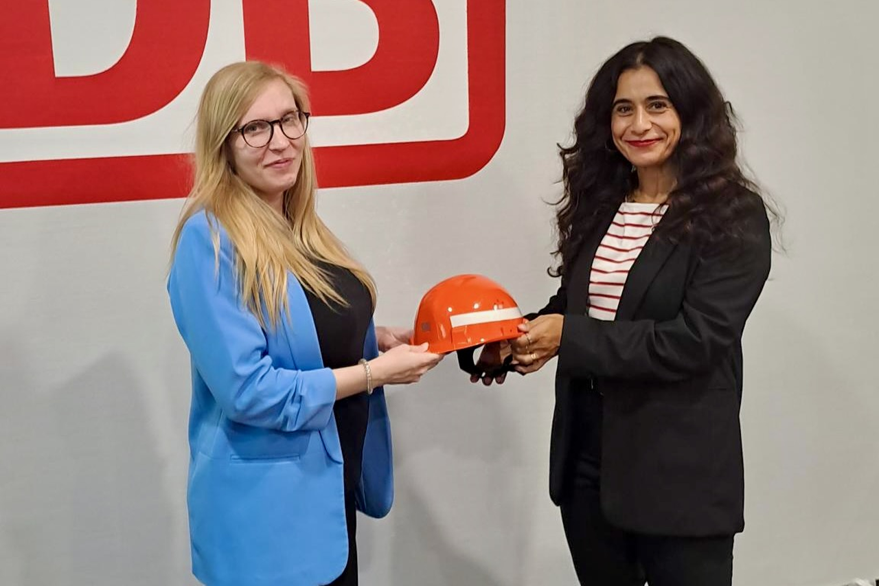 Zwei Personen stehen vor einer Wand mit großem DB-Logo. Eine Person überreicht einer anderen Person einen orangefarbenen Bauhelm.